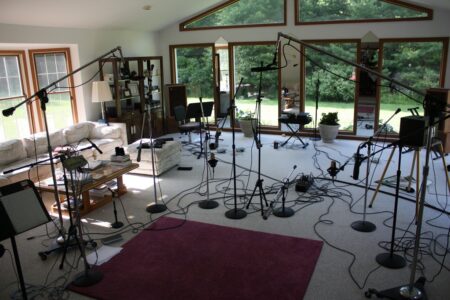 A recording session setup in a living room with multiple microphones and equipment on stands, surrounded by couches and large windows.
